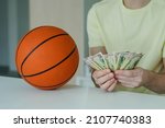 Small photo of Lucky man with basketball ball counting dollar banknotes. Close up of male hands count money cash payout after win at basketball betting. Concept of betting, gambling.