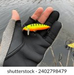 Small photo of Fisherman next to a small river holding a colourful bait in his hand. A fisherman holds a multicolored wobbler in his hands in close-up. The concept of catching predatory fish by spinning.