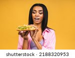 African American black beautiful young woman eating croissant or sandwich isolated over yellow background. Unhealthy junk food diet concept.