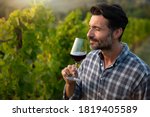 Authentic shot of happy successful male winemaker is tasting a flavor and checking red wine quality poured in transparent glass on vineyards background at sunset.