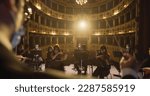 Small photo of Unfocused Photo for Background Usage: Back View of Professional Conductor Directing Symphony Orchestra with Performers on Classic Theatre with Curtain Stage During Music Concert. Out of Focus