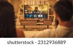 Small photo of Back View of Young Indian Couple Watching TV while Sitting on a Couch in the Living Room. Relaxed Evening for Enjoying Favourite Movies, TV Shows, Content and Ads Thank to Streaming Online Service