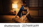 Small photo of Portrait of a Focused Adult Female Playing on a Traditional Japanese Toto Instrument. Musician Wearing a Beautiful Kimono, Performing a Classical Solo Indoors in a Dark Room with Lamp Light