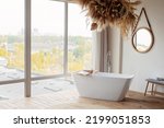 Comfortable bright bathroom with a boho-chic interior design, a free-standing white bath against the background of a panoramic window. hanging decor from dry grass. Soft selective focus.