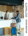 Small photo of Female Hotel Receptionist Assisting Businessman for Checking In. Business man with luggage talking with concierge on hotel reception with sneeze guard protection.