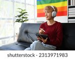 Satisfied young gay queer man in headphone and red sweater using digital tablet in living room with pride rainbow flag on background