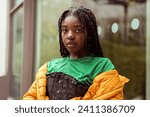 Small photo of Orange vibes - Diversity: Young black woman in her 20's with long braids on urban street style background wearing a vibrant orange jacket, green t-shirt and black corset. Fashion trend