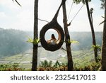 Small photo of A young woman tourist sitting in a bird nest, immersed in the breathtaking green landscape of Bali on a sunny day. Tourist doing jungle swing. Rice fields and forest in background. Indonesia.