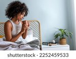 Young African American woman having morning coffee in bed. Multiracial latina female relaxing in bedroom having tea.