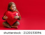 cute little girl sitting on the floor and hugging a red heart, on a red background. Happy valentine's day concept. Red heart shaped balloon