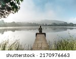 Calm misty morning meditation by pond.Sitting woman outdoors.Wellbeing and wellness soul concept.Spring foggy nature.Silence.Woman feeling freedom,enjoying vacation.No stress,calm mind,relax