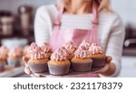 Small photo of Young caucasian woman holding cupcakes in the kitchen. Cakes cupcakes and sweet dessert pink girly concept. holding her homemade sweets housewife portrait Pastry chef confectioner