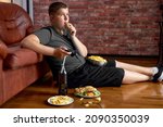 Fat teenager boy sitting on floor in living room, side view. Overweight obese caucasian child in casual clothes enjoy leading unhealthy lifestyle, eat junk food and play video games.