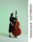 Small photo of Girl musician with long hair in black dress plays the double bass against the light green background.