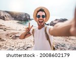 Small photo of Young man with backpack taking selfie portrait on a mountain - Smiling happy guy enjoying summer holidays at the beach - Millennial showing victory hands symbol to the camera - Youth and journey