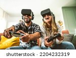 Small photo of Happy friends sitting on sofa playing video games with virtual reality glasses - Cheerful couple having fun with modern technology videogames - Hobby and tech concept