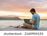 Young digital nomad man sitting on wooden pier at sea working on internet remotely at sunset - Traveling with a computer - Online dream job concept - Selective Focus
