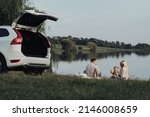 Happy Young Family Enjoying Picnic Time Outside City, Mother and Father with Their Daughter Sitting Outdoors Near Their SUV Car, Road Trip Concept