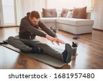 Small photo of Young ordinary man go in for sport at home. Freshman or beginner stretching with tow hands to one leg. Excercising alone in empty apartment. Try to keep fit and stay healthy with good body