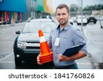 Male driving instructor standing with orange traffic cones and clipboard in his hands. Caucasian man working in driving school. Blurred gray car in background.