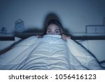 Woman watching a scary horror movie on tv late at night, she is frightened and hiding under the blanket
