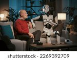 Senior man and female AI robot sitting on the couch at home and talking together, human-robot relationship concept