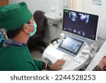 Small photo of Indonesian doktor observes the patient with a fetal detection device. An obstetrician wearing sterile one-off suit and scrub cap watching the screen of ultrasonographic diagnosis.