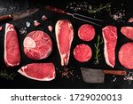 Cuts of meat, overhead flat lay shot on a dark background with salt, pepper, rosemary and knives