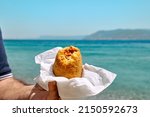 Small photo of Typical Sicilian street food on seaside background. Hand of man holding hot palatable arancina (deep fried rice balls with meat) in the beach. Take out food.