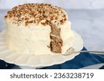 Three tier hummingbird or carrot cake with cream cheese frosting and pecan nuts or walnuts on a white cake stand with a slice being served with a cake lifter