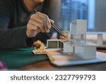 Small photo of Close-up hand of Architect students diligently make house model building samples with paper art architecture tools at night in their alone room.