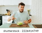Small photo of A young dieting man points his index finger at a bowl of salad in front of him, asking Is this supposed to be eaten Green salad healthy food concept. Sits in his kitchen. Looking at camera.
