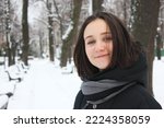 An attractive young brunette woman stands in snow-covered winter park smiles looking at camera. Portrait of pretty female girl. Path covered with white snow goes into distance. Wooden benches in a row