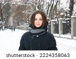 An attractive young brunette woman stands in snow-covered winter park smiles looking at a camera. Portrait of a beautiful female girl. A path covered with white snow goes into distance. Cold weather