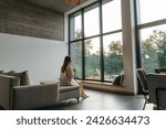 Woman in a country house or hotel with panoramic windows in pine forest sitting on the bed and enjoys beautiful view with light sun in the morning. Good morning and recreation on nature concept 