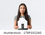 Small photo of Insurance, loan, real estate and family concept. Indecisive and confused asian woman thinking about buying or renting home, holding paper house and looking away thoughtful, white background
