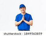 Small photo of Friendly-looking handsome asian delivery man in blue uniform, holding hands in plead, palms together to say thank you, cherish and value customers. Courier looking happy and grateful
