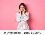 Cute and shy asian girl blushing, touching cheeks and looking left at copy space silly, standing against pink background