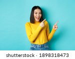 Small photo of Shopping concept. Beautiful chinese girl in yellow sweater pointing fingers at upper right corner logo banner, smiling amused, standing over blue background