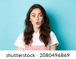 Small photo of Close up portrait of excited stylish girl with curly hairstyle and beauty make up, pucker lips intrigued and stare at camera with surprised face, standing over blue background