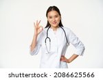 Small photo of Very good. Smiling confident asian female doctor, showing okay, ok sign in approval, confirm smth, saying yes, give approval, standing in medical uniform, white background