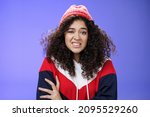 Small photo of Woman with curly hair in winter beanie feeling uncomfortable and discomfort clenching teeth and frowning intense as hugging herself insecure and awkward, unwilling to say cruel rejection, feel awkward