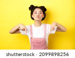 Small photo of Sassy asian teen girl pucker lips and pointing fingers down, tell to look at bottom advertisement, standing with glamour makeup and summer clothes on yellow background