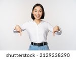 Small photo of Portrait of young asian woman pointing fingers down and smiling, showing banner, click on link below gesture, inviting people to follow, standing over white background