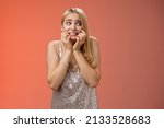 Small photo of Frightened insecure timid young blond woman afraid feel scared trembling fear stooping biting nails pop eyes right standing horrified red background wear silver evening dress terrified