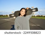 Leisure and people. Happy asian woman standing with longboard, cruising on an empty road in countryside. Skater girl holds her skateboard and smiles at camera.