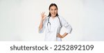 Small photo of Confident female doctor, asian physician in medical uniform and stethoscope, showing okay sign and nod pleased, praising, recommending smth good, white background.