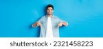 Small photo of Handsome smiling adult man introduce product, pointing fingers down at promotion, standing against blue background.