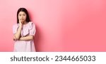 Small photo of Bored asian teen girl look indifferent at camera, lean face on hand in skeptical pose, standing reluctant against pink background.