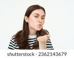 Small photo of Portrait of cute girl sending you, blowing air kiss, holding palm near puckered lips, smiling, standing over white background.